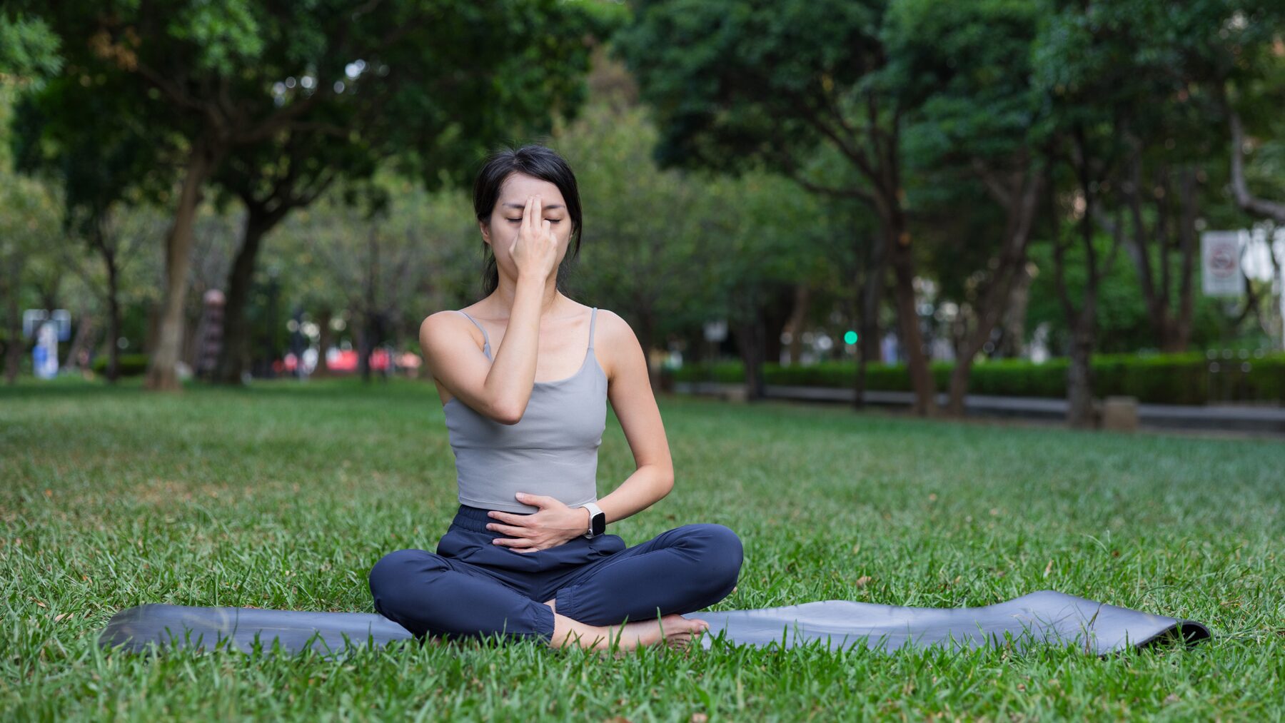 A woman practising alternate nostril breathing on a mat in a green park